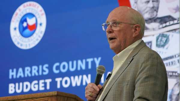 Speaker presenting at a Harris County budget town hall meeting, standing behind a wooden podium with a microphone and a presentation screen displaying county budget information.