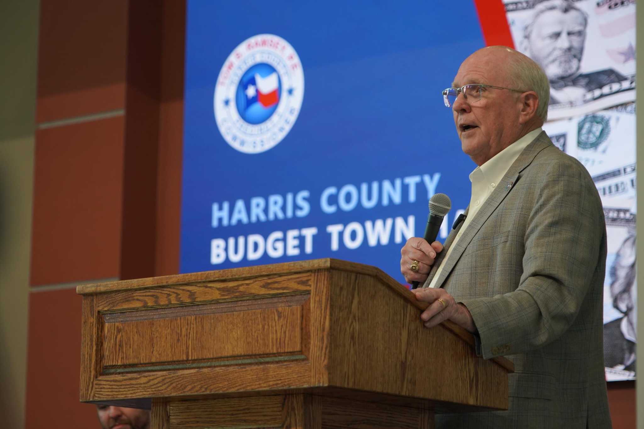 Speaker presenting at a Harris County budget town hall meeting, standing behind a wooden podium with a microphone and a presentation screen displaying county budget information.